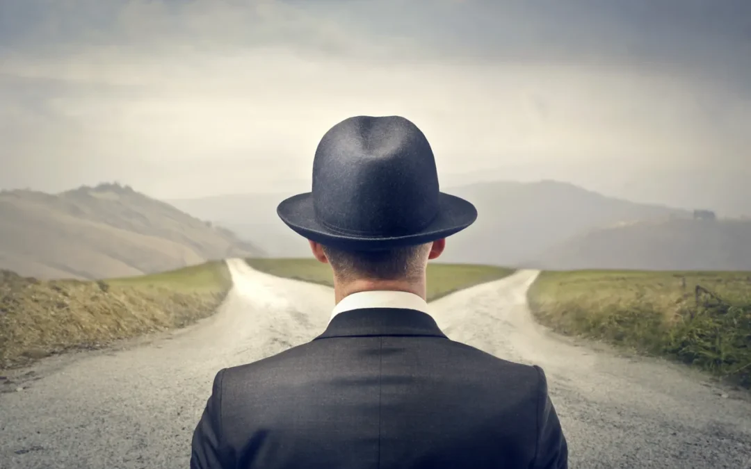 Man in a black suit and a black hat standing in front of two diverging roads leading into the hills representing choice